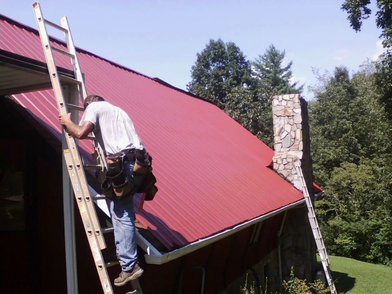 Roofer on a ladder beside a red metal roof and stone chimney — Soffit & Fascia Repair by Roofing Brothers in North Little Rock